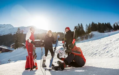 Grupo de amigos ri e se diverte na neve, enquanto carrega equipamentos de ski