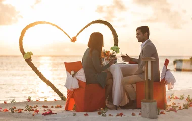Casal em almoço romântico à beira da praia durante o pôr do sol com decoração em formato de coração e pétalas vermelhas no chão