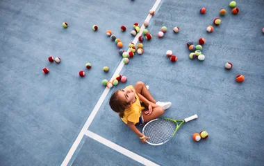 Criança sentada na quadra de tennis em meio às bolas.