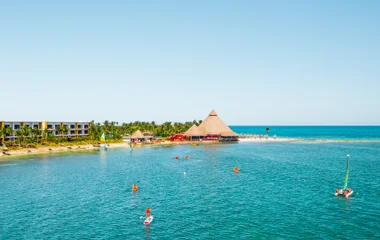 Mulher senta na areia à beira mar e toma banho de sol enquanto homem ao seu lado toma sol dentro de um caiaque amarelo, também à beira mar