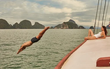 Casal em barco e homem pulando no mar.
