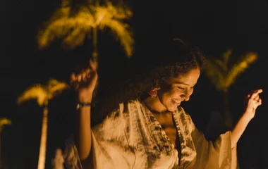 Uma pessoa dançando na festa com os braços para cima em um ambiente escuro com uma palmeira atrás