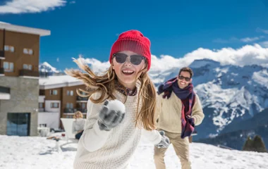 Menina ri e se diverte com bola de neve em sua mão, seu pai é visto logo atrás também sorrindo em meio a paisagem invernal.