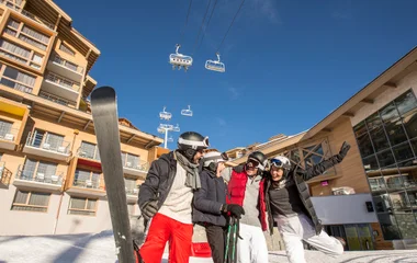 Grupo de amigos ri e se diverte na neve, carregando equipamentos de esqui, com o resort Club Med Val Thorens atrás.
