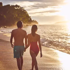 Casal de costas com roupas de banho, na areia à beira da praia e com por dol sol ao fundo
