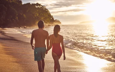 Casal de costas com roupas de banho, na areia à beira da praia e com por dol sol ao fundo