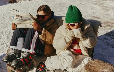 Criança toma chocolate quente sentada na neve e usa gorro verde e óculos escuros