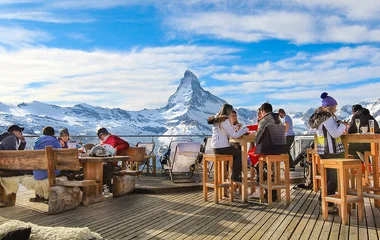 Pessoas sentadas num restaurante a céu aberto com a montanha nevada Zermatt ao fundo, dia ensolarado.