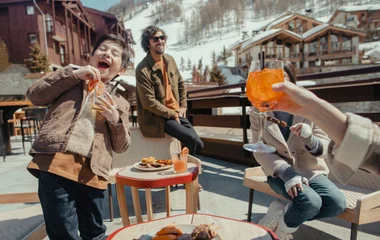 Família em frente aos chalés com a neve ao fundo, felizes tomando drinks e comendo snacks