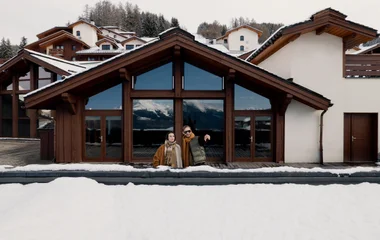 casal em frente a uma varanda com telhado coberto de neve observando a paisagem