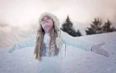 Menina vestida toda de branco com capuz de pele de braços aberto na neve.