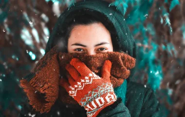 Mulher cobre a boca com cachecol marrom e está usando luvas e capuz, enquanto neva