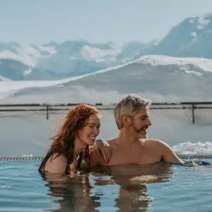 Casal em piscina aquecida com paisagem de neve ao fundo.