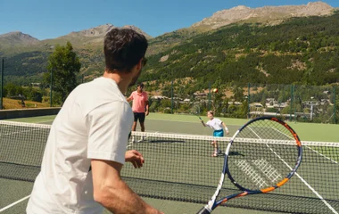Um adulto de costa jogando tênis contra outro adulto e uma crianças no meio de um set em uma quadra verde tradicional tradicional do esporte com montanhas verdes ao fundo